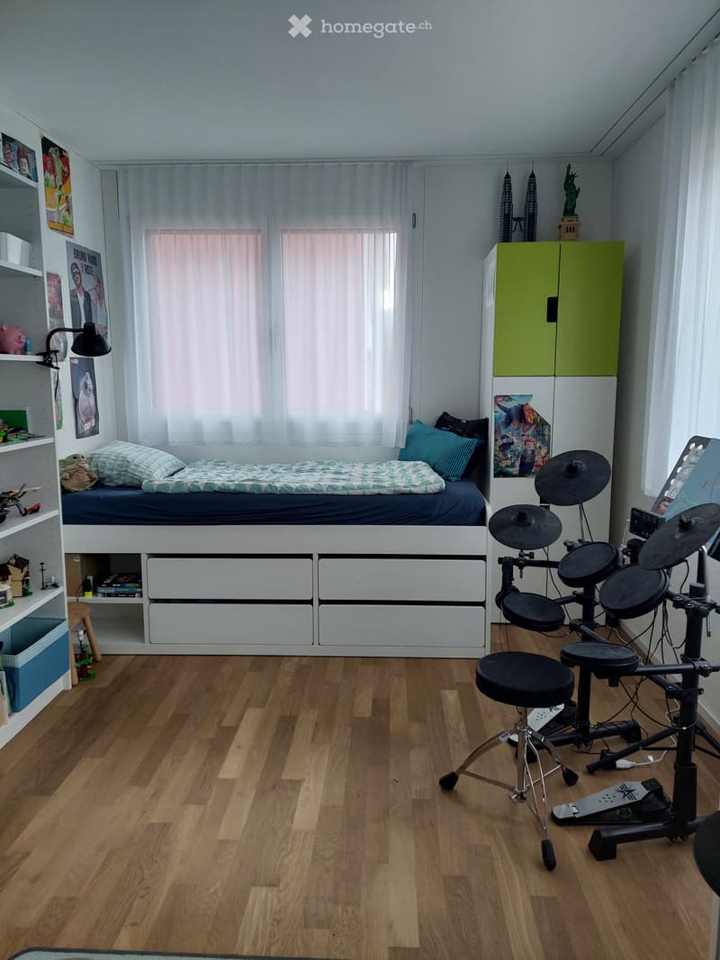 A well-lit bedroom with a lofted twin-sized bed, a drum set, a stool, a shelf with books and toys, and a window with white curtains.