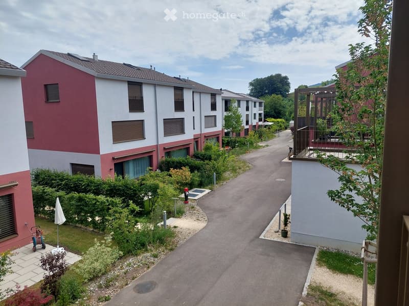 Residential complex with modern two-story buildings, red and white facades, balconies, and landscaped gardens.