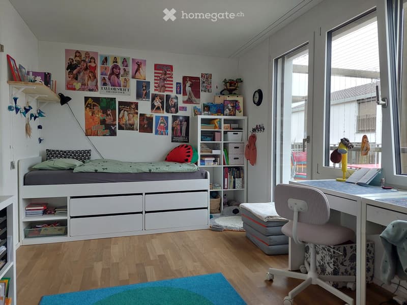 A modern, well-lit bedroom with a lofted twin-sized bed, built-in storage drawers, a desk with a chair, a bookshelf, and various decorative posters and artwork on the walls.