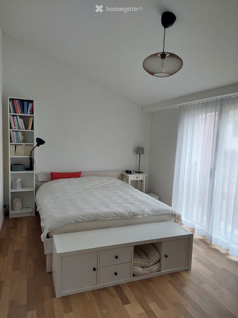 Modern bedroom with a large bed, nightstand, bookshelf, and a bench at the foot of the bed. The room features wooden flooring, white walls, and large windows with sheer curtains.