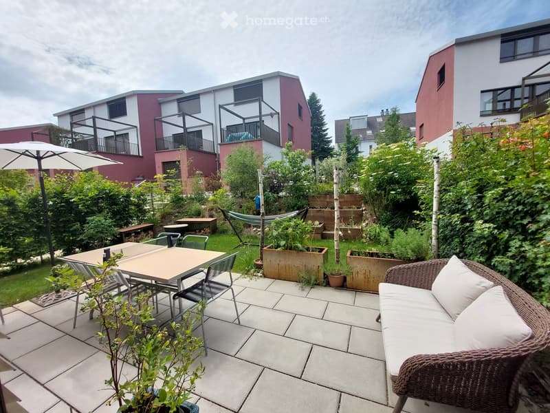 A well-maintained garden patio with a wicker sofa, dining table with chairs, a hammock, and potted plants. The area is surrounded by greenery and features modern residential buildings in the background.