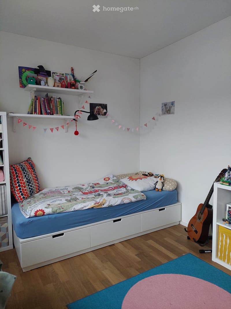 A cozy children's bedroom with a single bed, colorful bedding, a guitar, shelves with books and toys, and a rug on the floor.