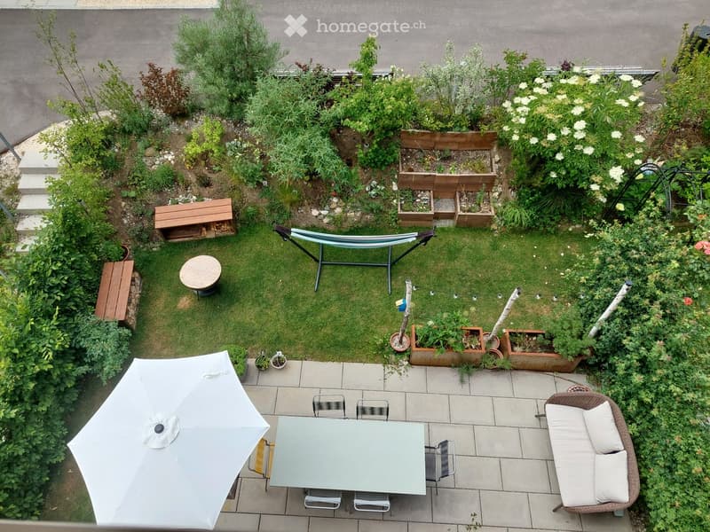 A well-maintained garden with a small seating area, including a table, chairs, umbrella, and a wicker chair with cushions. There is a hammock, a wooden bench, and various potted plants and flower beds.