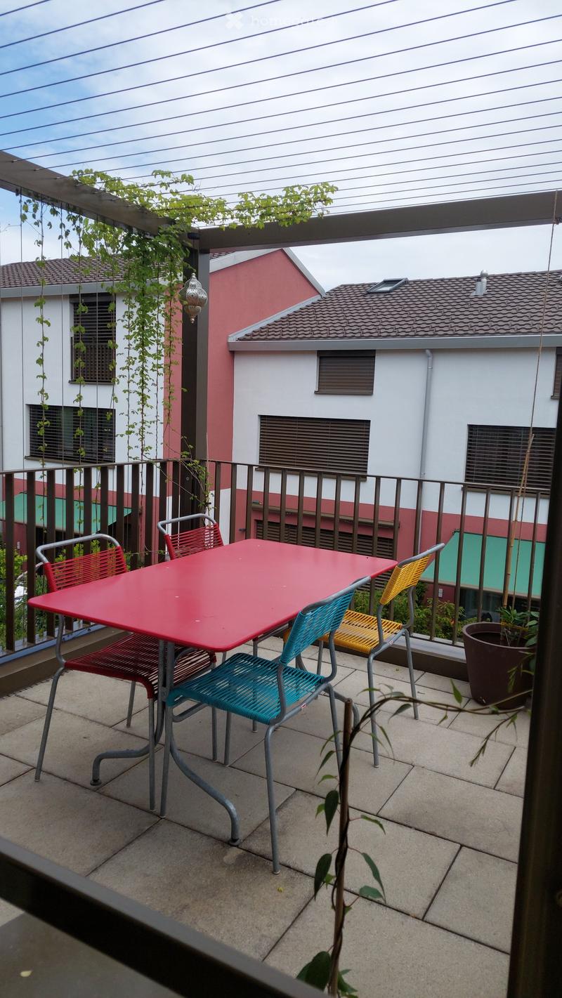 Balcony with a red table and colorful chairs, surrounded by a metal railing and potted plants.