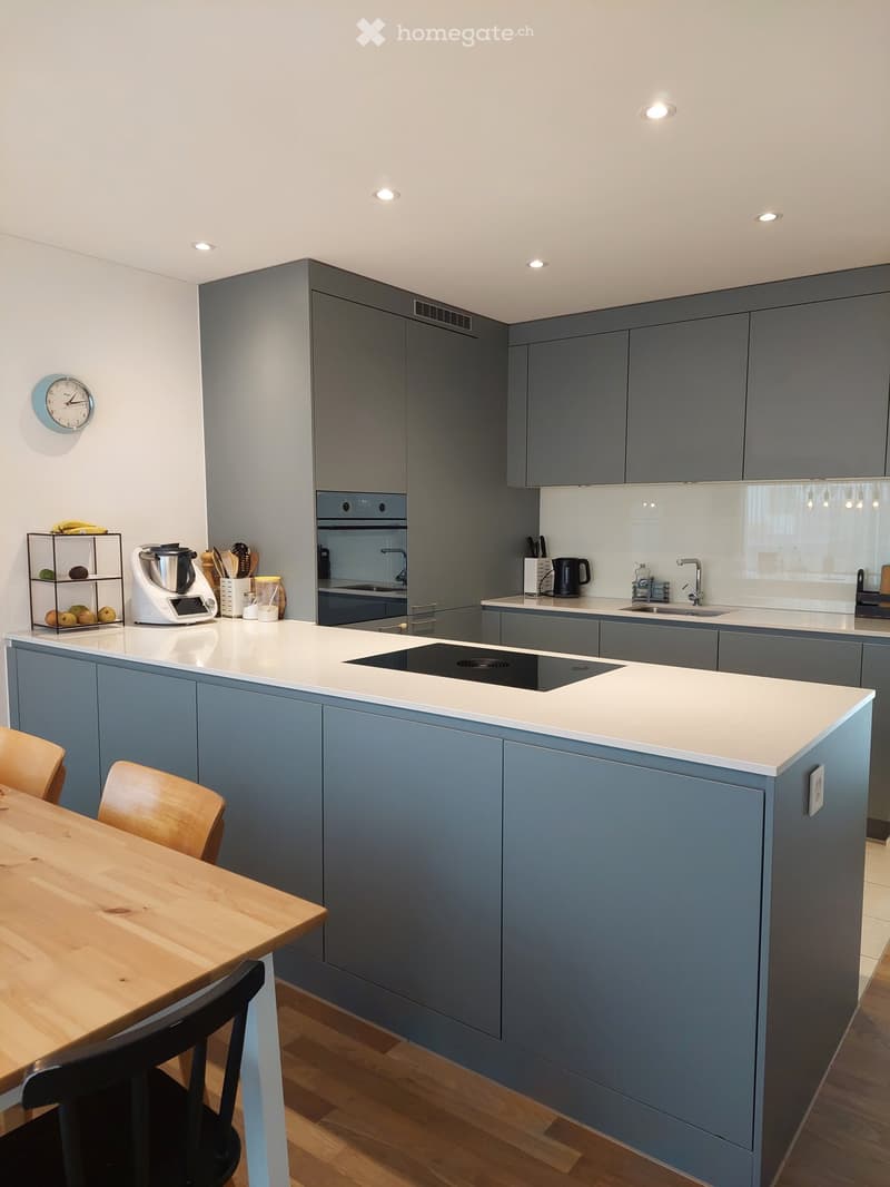 Modern kitchen with grey cabinets, white countertops, and a central island with an induction cooktop. Includes a wooden dining table with chairs, a blender, and a sink with a faucet.