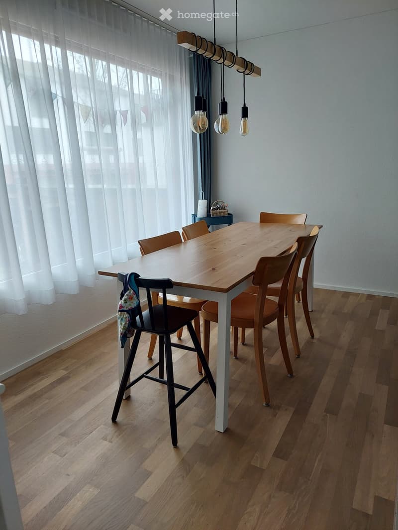 Dining area with a wooden table, multiple chairs, a stool, and a hanging light fixture. The room features wooden flooring and large windows with white curtains.