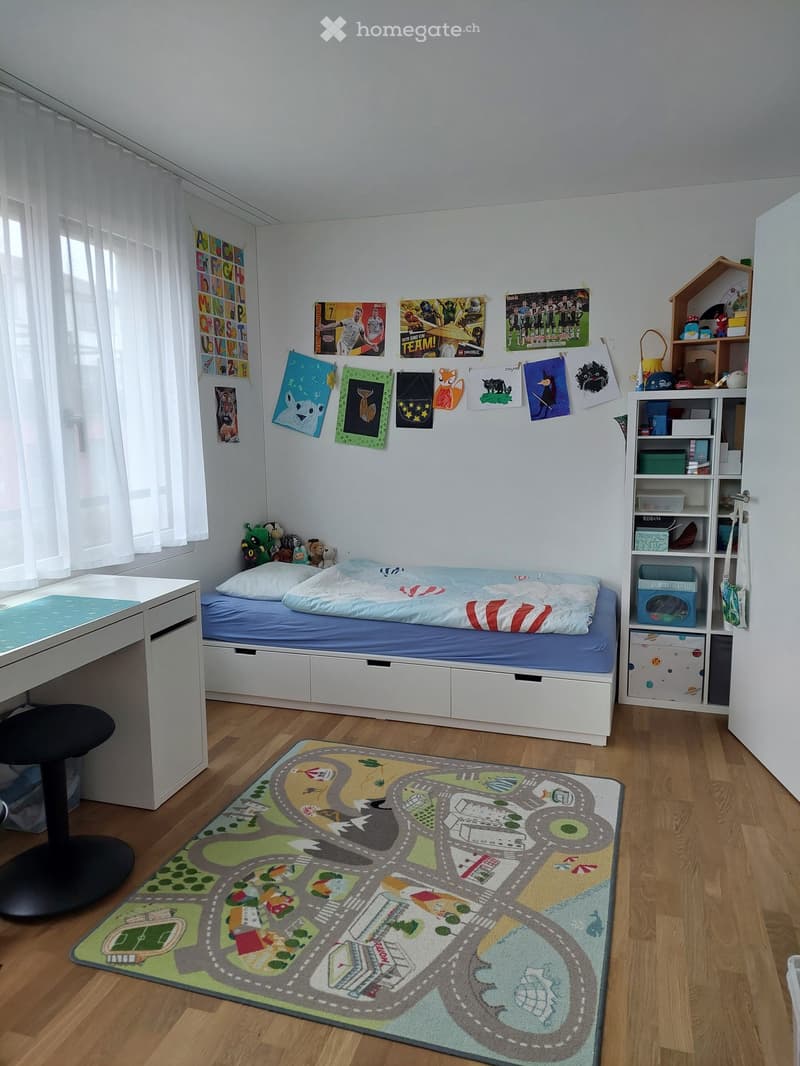 A well-lit bedroom with a single bed, desk, and shelf. The room features a colorful road-themed rug, various posters, and a small toy house on the shelf.