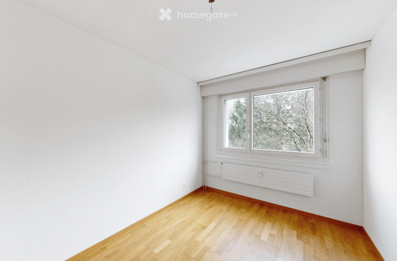 Completely empty room with white walls, wooden floor, and a large window with a view of trees.
