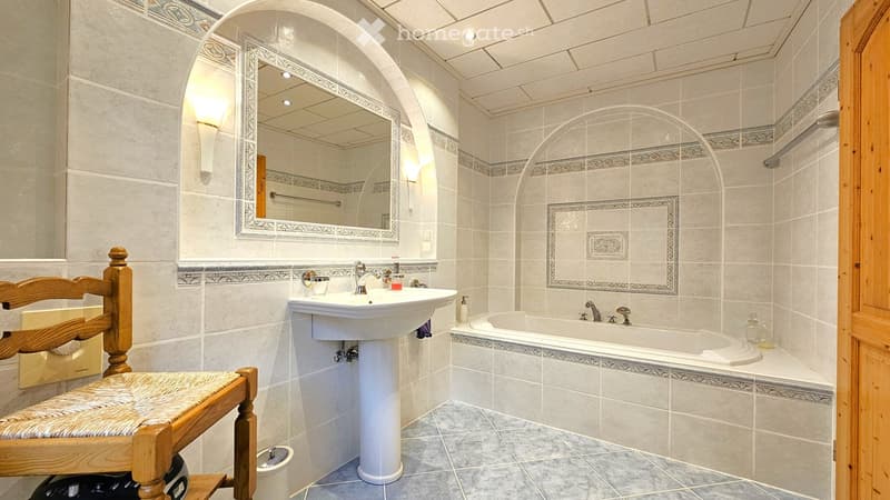 Elegant bathroom with a white sink, bathtub, wooden stool, and decorative mirror. Light-colored tiles and warm lighting create a cozy atmosphere.