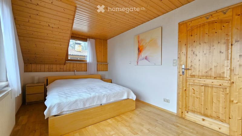 Cozy wooden bedroom with a large bed, nightstand, and abstract painting on the wall. The room features wooden floors and a slanted wooden ceiling.