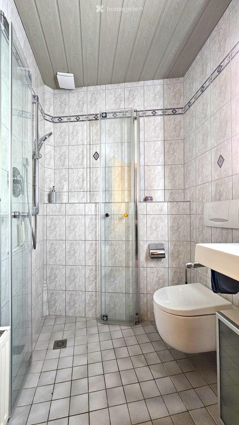 Modern bathroom with a toilet, sink, and glass-enclosed shower. The walls and floor are tiled in a light, neutral color with decorative borders. A towel is draped over the edge of the sink.