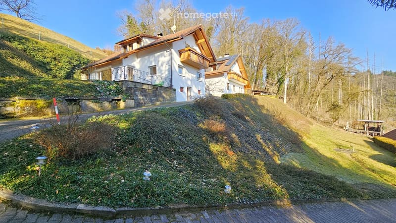 Two-story house with white walls, wooden balconies, and a sloped roof, situated on a hillside with surrounding trees and a clear blue sky.