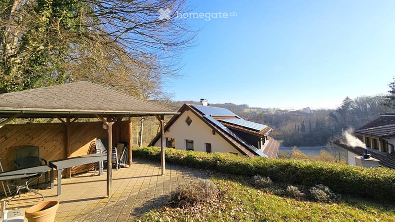 A wooden patio with a table and chairs, surrounded by a well-maintained garden and a modern house with solar panels on the roof.