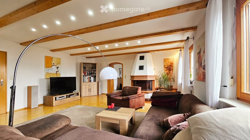 Cozy living room with exposed wooden beams, a fireplace, a large sectional sofa, an armchair, a coffee table, a bookshelf, and a television. The room features large windows with white curtains and a floor lamp with a white shade.