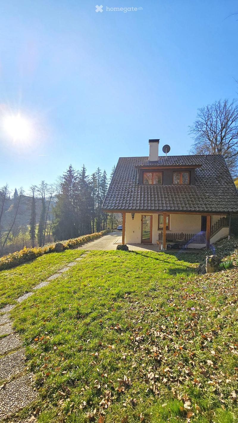 Two-story house with a tiled roof, chimney, and satellite dish, situated on a grassy slope with a wooden bench and small patio area.