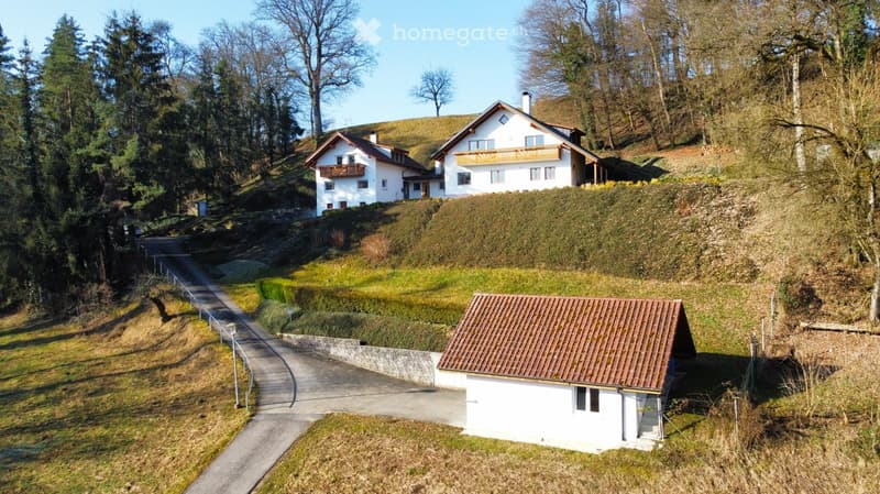 Aerial view of a residential area with three houses on a hillside, surrounded by trees and greenery. The houses have white walls, pitched roofs, and balconies.