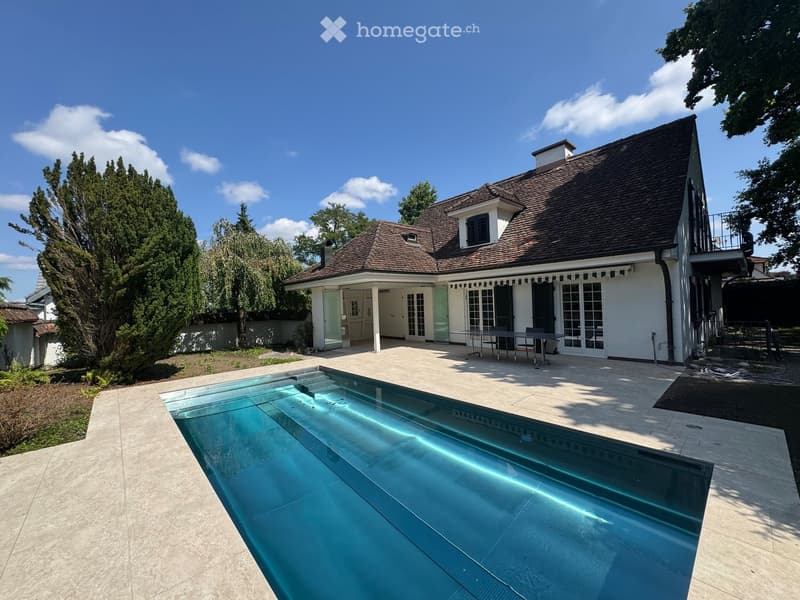 Two-story house with a tiled roof, white exterior, and a large swimming pool in the foreground. The house features a covered patio area with outdoor furniture and is surrounded by trees and greenery.