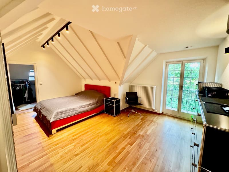 Bright attic bedroom with wooden flooring, slanted ceilings, and modern furnishings including a red bed, black nightstand, and a desk area with a glass partition.