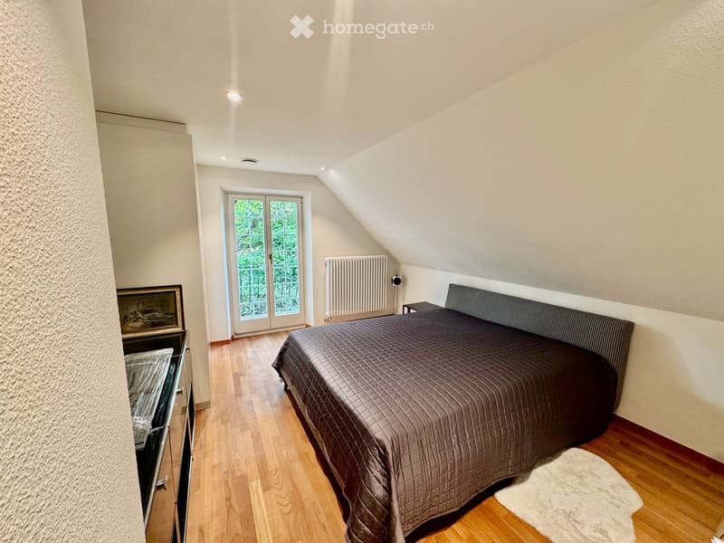 Cozy bedroom with wooden flooring, slanted ceiling, large window with glass door leading outside, bed with dark bedding, nightstand, radiator, and picture frame on wall.