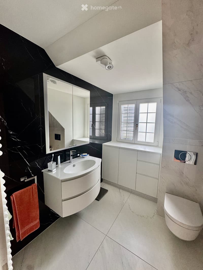 Modern bathroom with black marble walls, white sink, mirror, and toilet. Features a sleek vanity cabinet and a window with blinds.