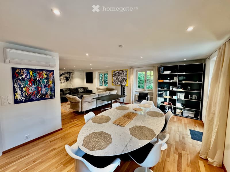 A modern living and dining area with a round marble dining table, white chairs, a sofa, a shelf, and a TV screen.