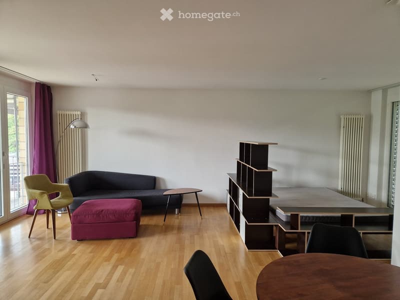 Modern living room with wooden flooring, a dark grey sofa, a purple ottoman, a yellow armchair, a small round table, a tall shelving unit, and a dining table with chairs.
