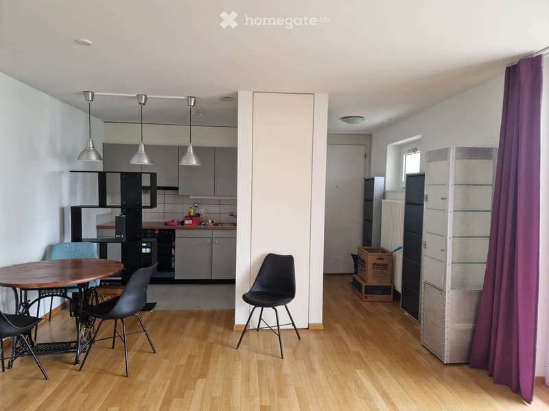A modern, open-plan living and dining area with a wooden floor, a round wooden dining table with four black chairs, a purple curtain, a kitchen area with grey cabinets, a sink, an oven, and a refrigerator, and a radiator in the background.