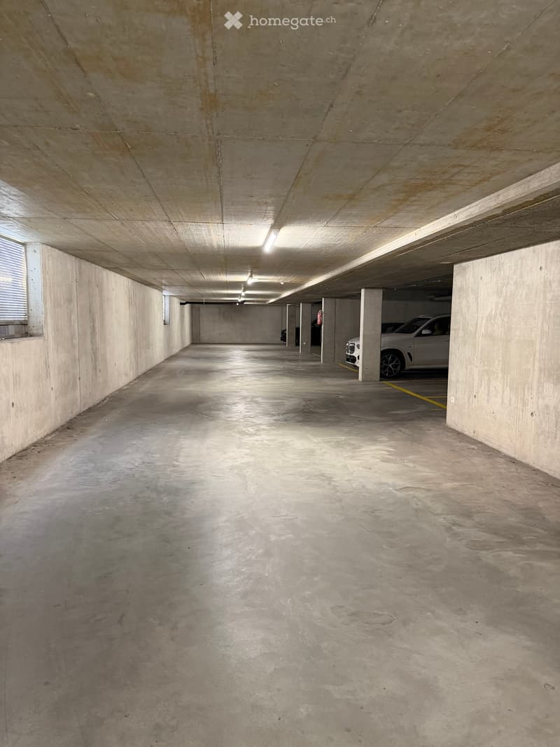 Underground parking area with concrete walls and ceiling, a few parked cars, and fluorescent lighting.