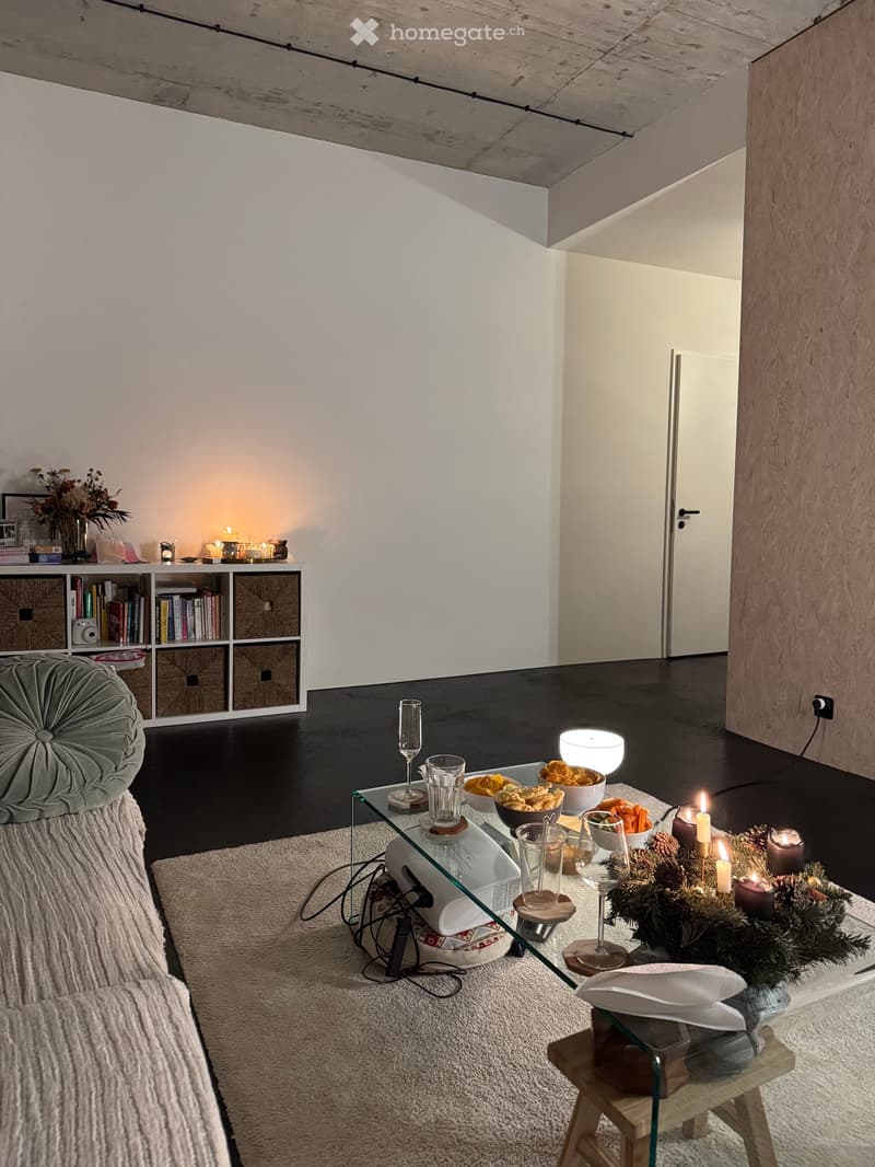 Modern living room with a dark wooden floor, white walls, and a high exposed wooden ceiling. Features a grey sofa, glass coffee table with decorative items, bookshelf, and a small wooden stool.