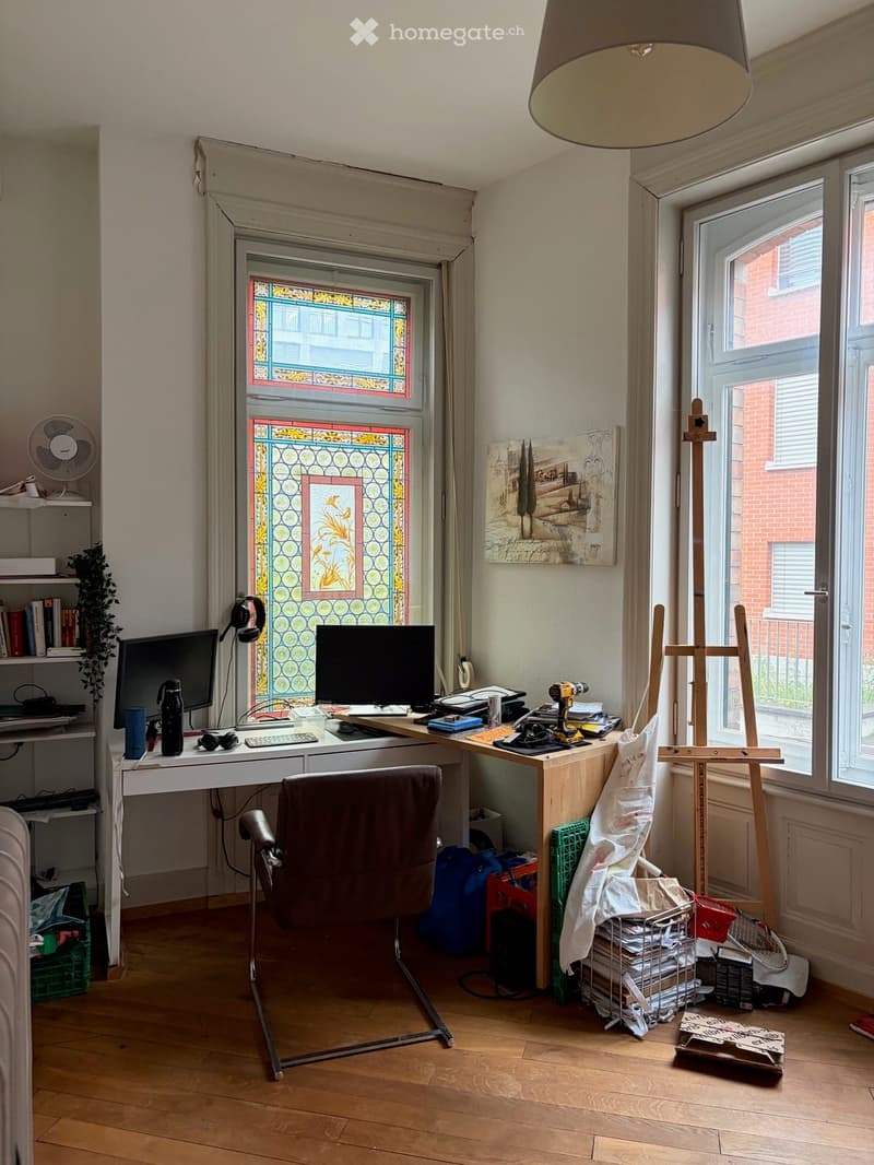 A well-lit room with wooden flooring, a large window with stained glass, a desk with a computer setup, a chair, a fan, a painting on the wall, and an easel with art supplies.