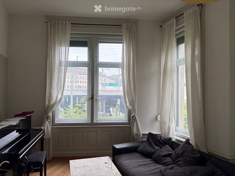 A cozy living room with a dark grey sofa, a white coffee table, a black piano, and a small table with a cup on it. The room has large windows with white curtains, wooden flooring, and a view of the street outside.