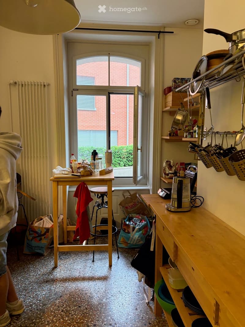 The image shows a small, well-lit kitchen with a wooden table, a stool, a coffee maker, shelves with pots and pans, and a window leading to a balcony. The kitchen features a tiled floor and a variety of kitchen items on the counter and shelves.