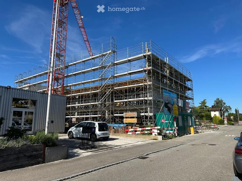 Multi-story building under construction with scaffolding, a crane, and a parking area with a white car.