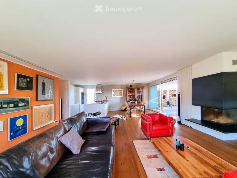 Modern living room with a black leather sofa, red armchair, wooden coffee table, fireplace, and an open kitchen area with a dining table and chairs.