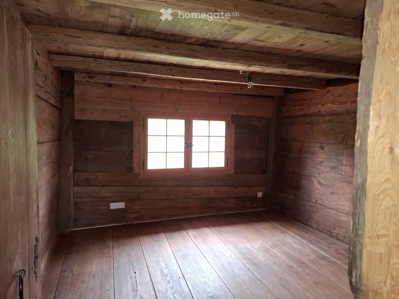 Empty wooden room with exposed beams, wooden floor, and a single window with multiple panes.