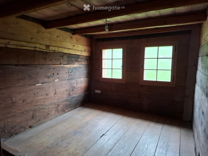 Empty wooden room with exposed wooden walls and ceiling, two windows, and a single light fixture.