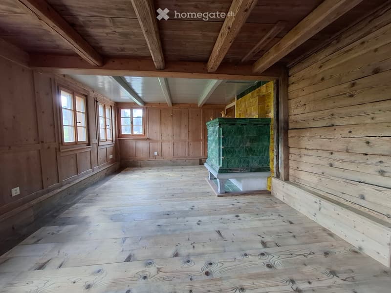 An empty wooden room with exposed beams, wooden walls, and a green chest in the center. The room has large windows and a wooden floor.