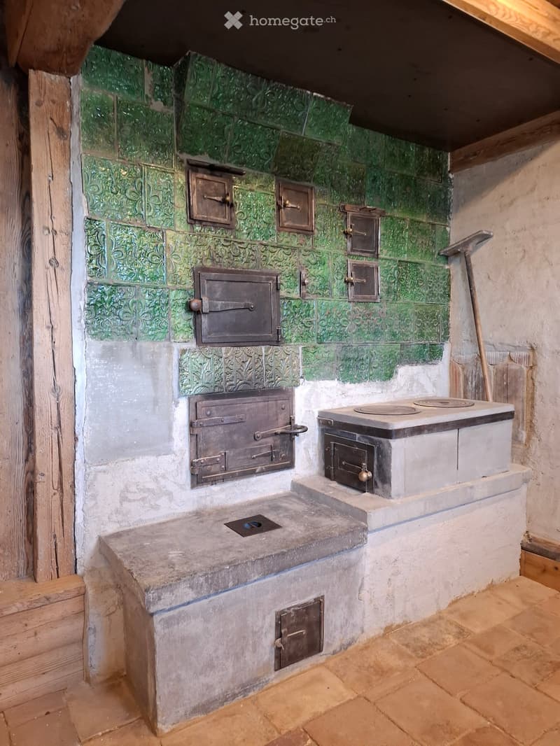 Traditional kitchen with a stone stove and multiple ovens, green decorative tiles on the wall, and a wooden broom leaning against the wall.