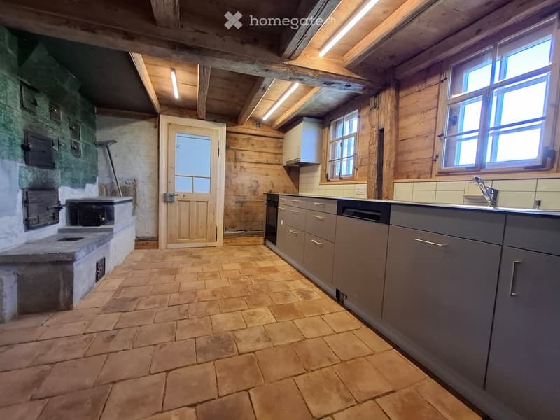 Modern kitchen with grey cabinets, stone tiled floor, and a stone fireplace on the left side.