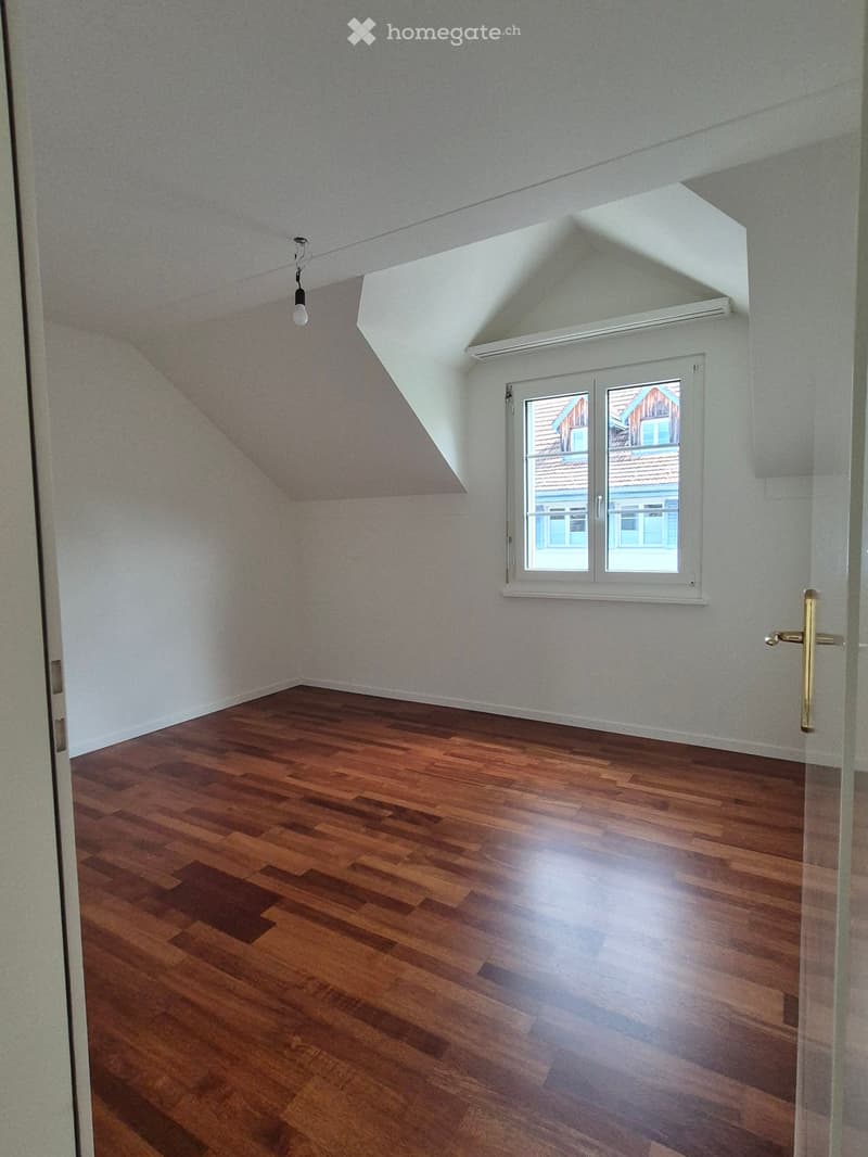 Empty room with wooden flooring, white walls, and a window with a view of neighboring houses.