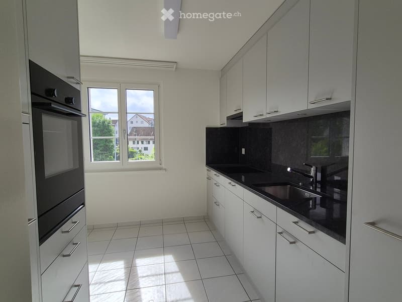 Modern kitchen with white cabinets, black countertops, a stainless steel sink, and an oven. Natural light streams in from a window showing a view of the street and neighboring houses.