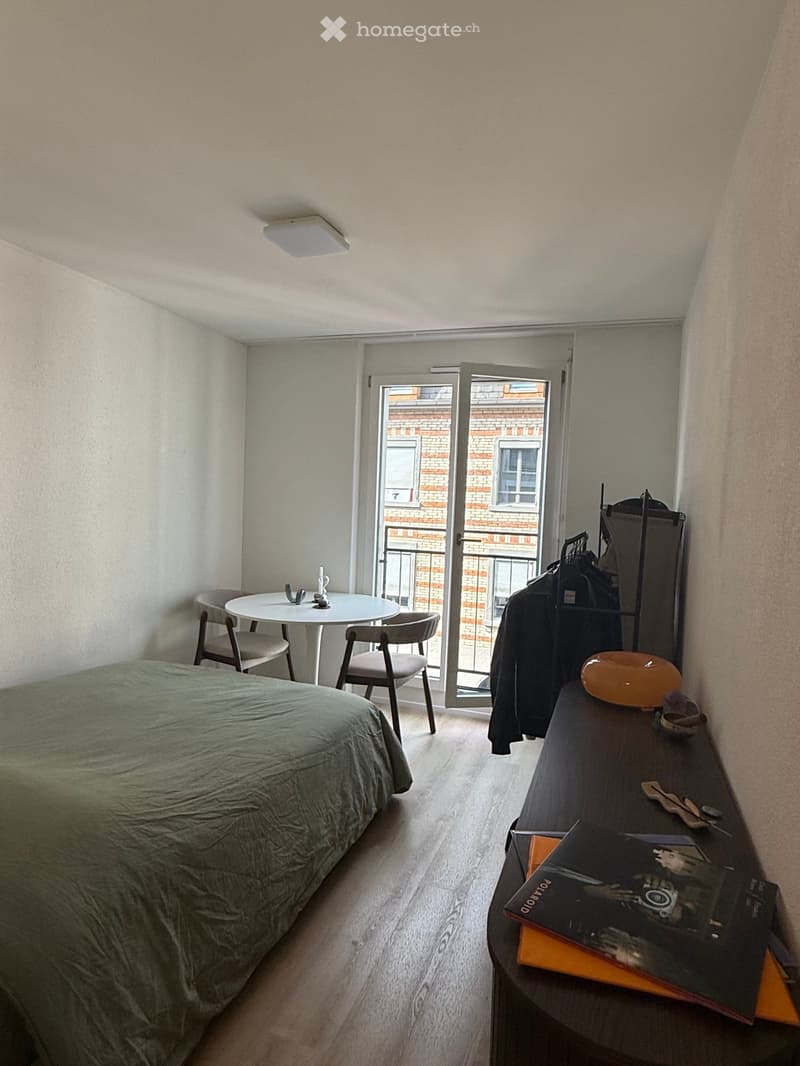 Cozy bedroom with a bed, small round table with chairs, wardrobe, and books on a side table. The room has wooden flooring and a sliding glass door leading to a balcony.