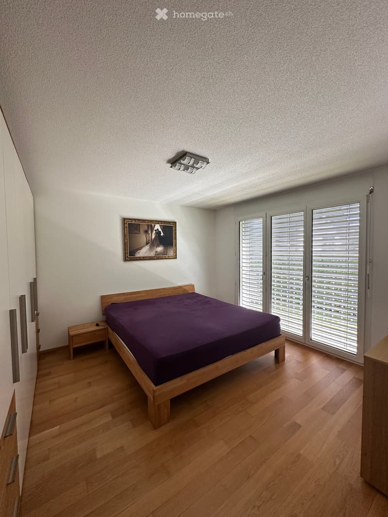 Modern bedroom with wooden flooring, a wooden bed with dark purple bedding, a nightstand, and a framed picture on the wall. Large windows with blinds provide natural light.