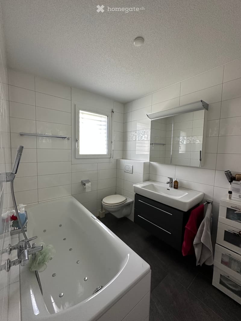 Modern bathroom with a bathtub, toilet, sink, mirror, and towel rack. Features tiled walls and a window for natural light.
