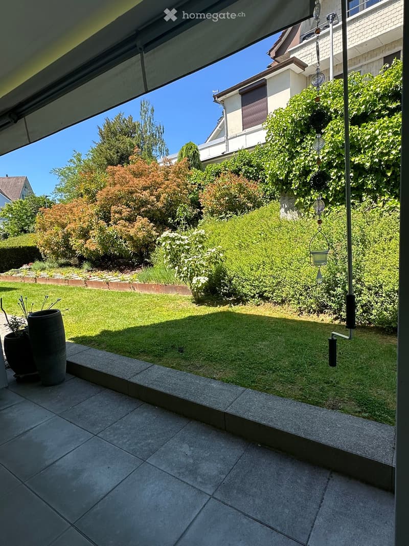Well-maintained garden with green lawn, various bushes, and trees, visible from a covered patio area with tiled flooring.