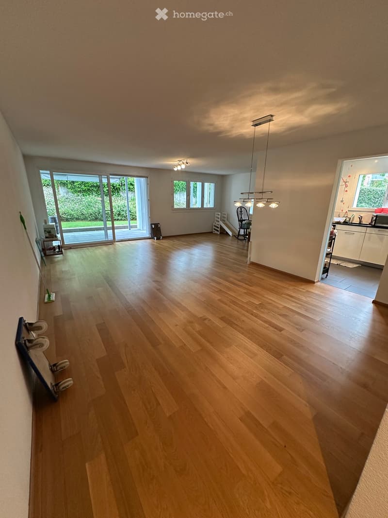 Empty room with light wooden flooring, large sliding glass doors leading to a garden, and visible kitchen area in the background.
