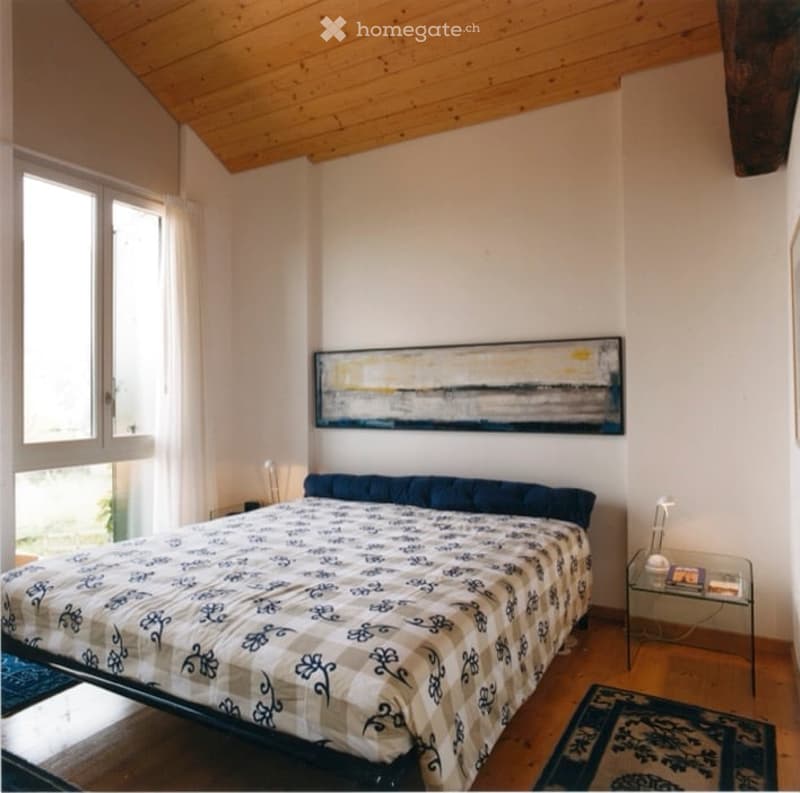 Cozy bedroom with wooden floors, a large bed with patterned bedding, a nightstand, a painting on the wall, and a small table with a chair.