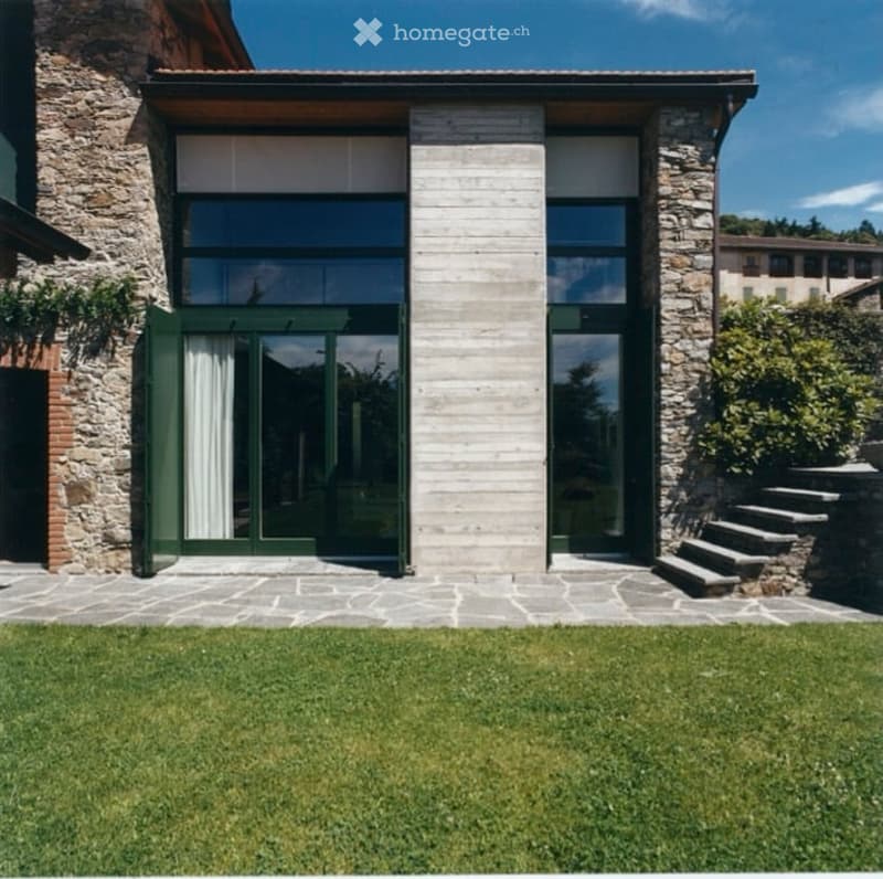 Modern house with stone and wood facade, large glass windows, green shutters, and a well-maintained lawn.
