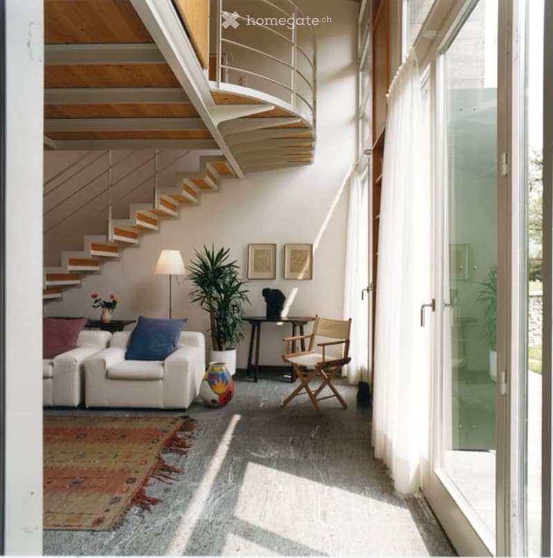 Modern living room with white sofa, wooden chair, small table, floor lamp, plant, and artwork on the wall. Features a curved staircase in the background and large windows allowing natural light.
