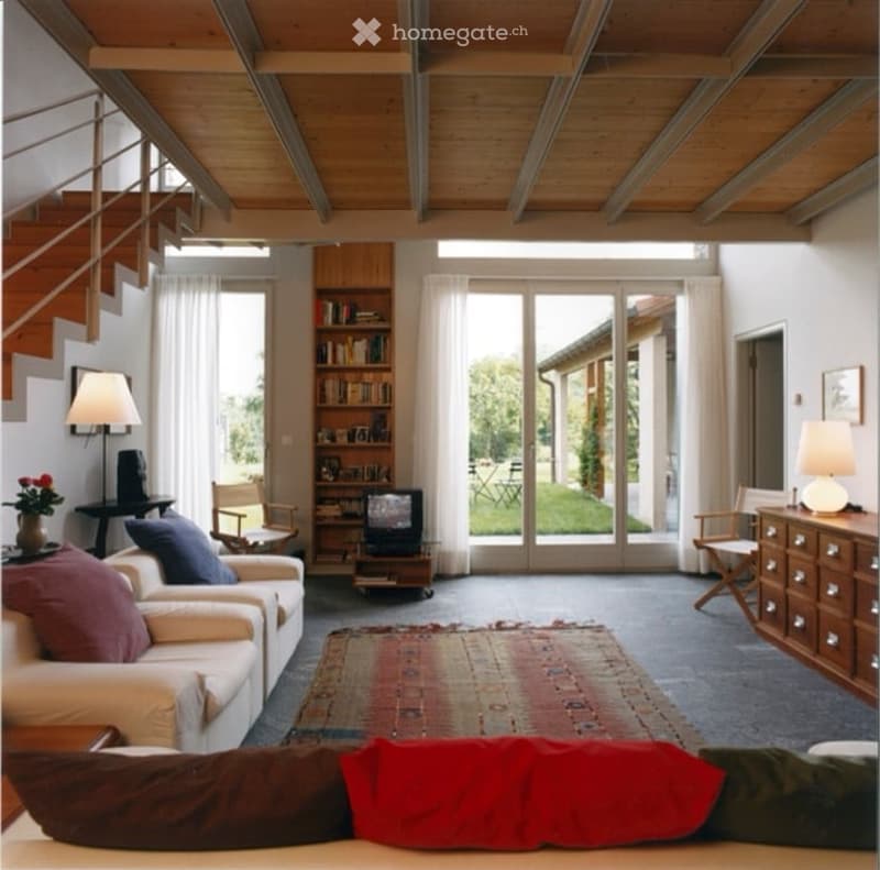 Bright living room with wooden ceiling beams, large windows, and sliding glass doors leading to a patio. Features a cream-colored sofa, wooden chairs, a small table with a lamp, a television on a stand, and a bookshelf with books and decorative items.